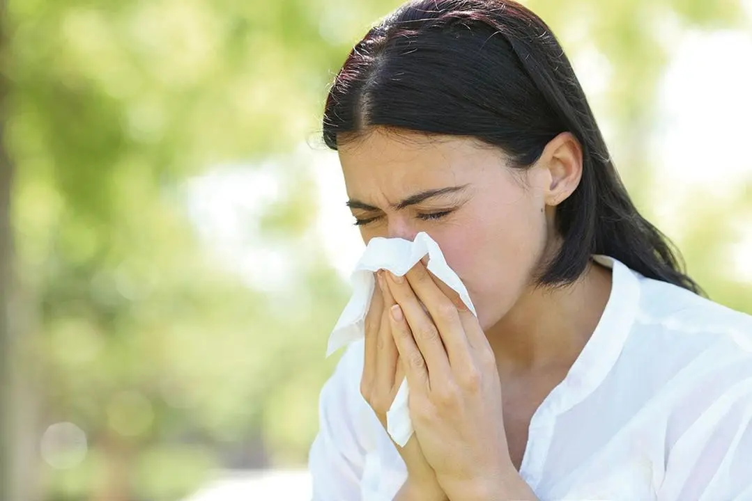 Ill woman sneezing covering mouth with a wipe standing in a park with a green background