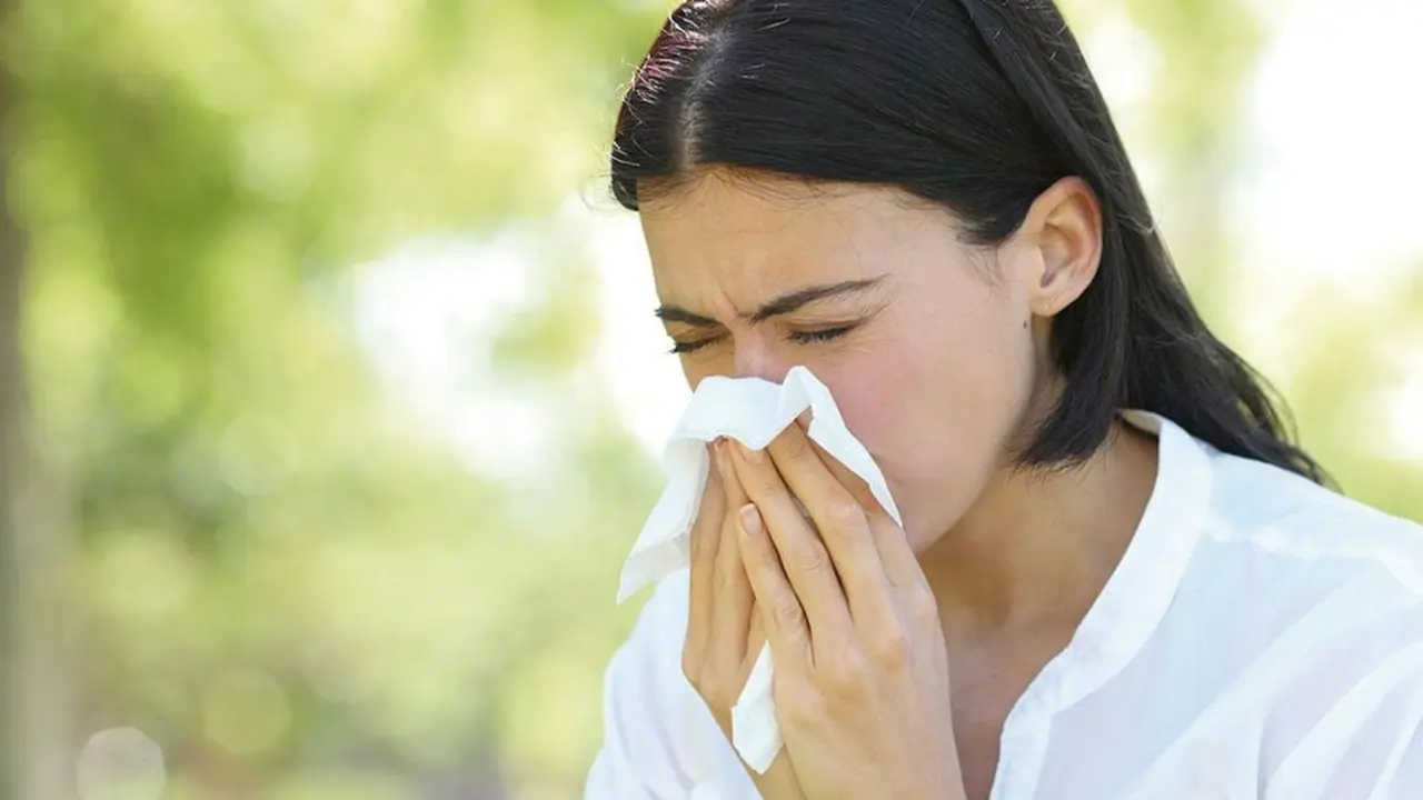 Ill woman sneezing covering mouth with a wipe standing in a park with a green background