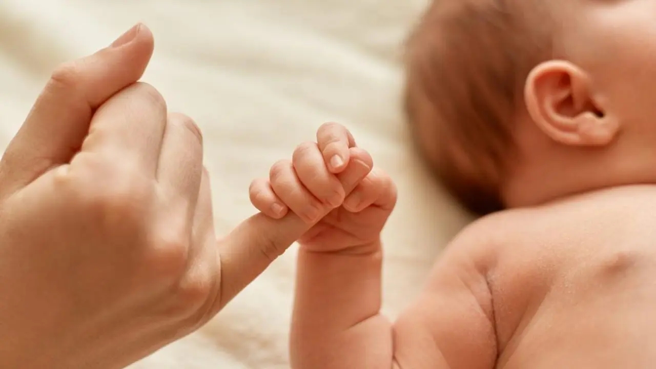 Faceless mother with naked baby, infant holding mommy's finger, mum spending time with her tiny child on light background.