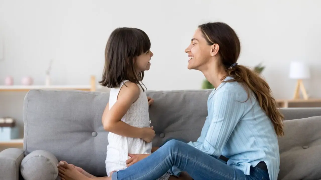 Attractive woman and little girl sitting on comfortable couch at home. Young mother talking communicates with small adorable daughter. Best friends happy motherhood weekend together with kid concept