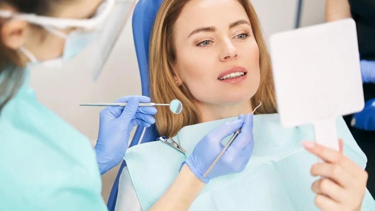 Female person lying in dental chair and looking in the mirror while dentist holding dental tools